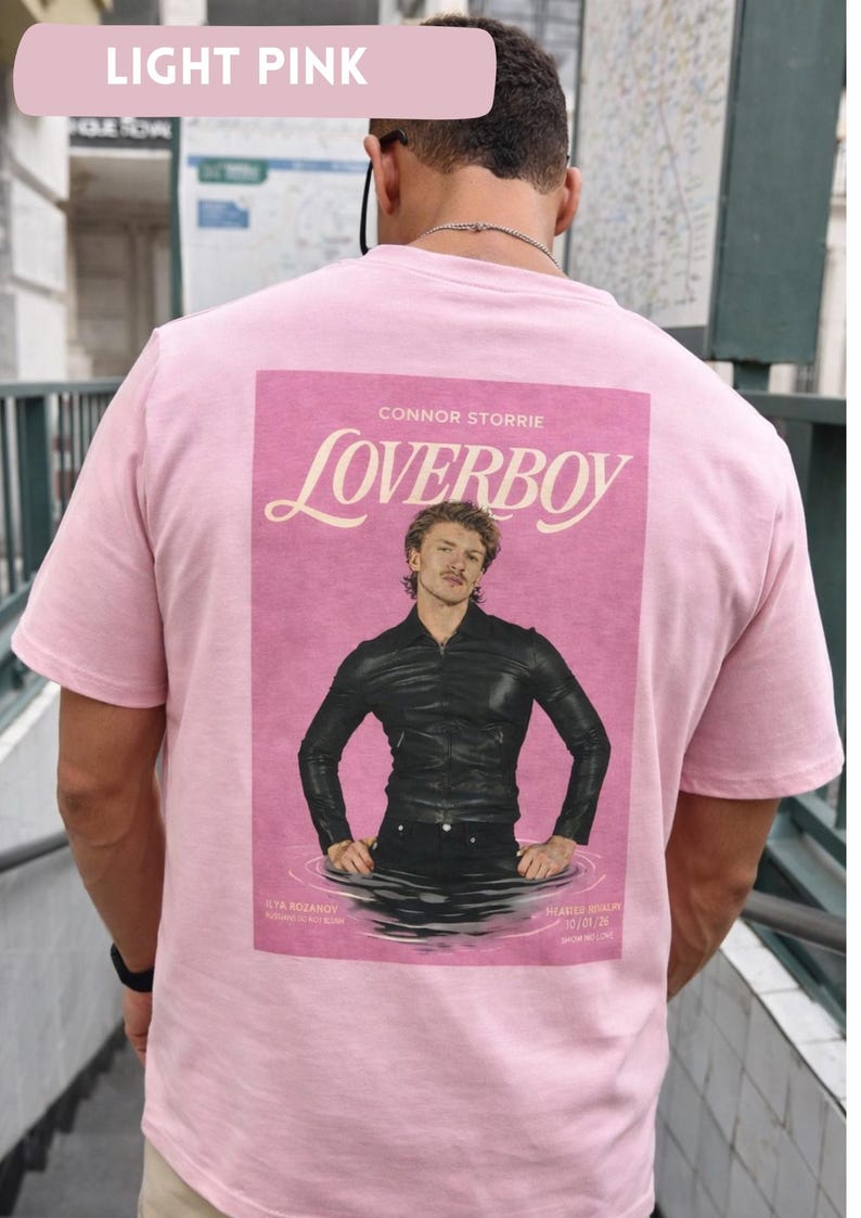 May include: Light pink t-shirt featuring a graphic of a man in a black leather jacket with the word "Loverboy" above the image. The text "CONNOR STORRIE" is above the word "Loverboy". The shirt is light pink.