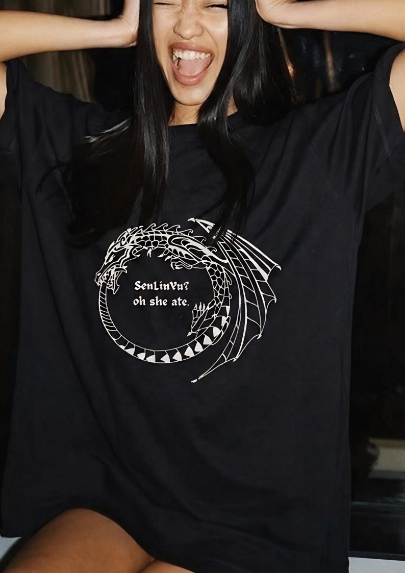 Op de afbeelding: Zwart T-shirt met een witte drakenafbeelding en de tekst "SenLinVu? oh she ate." Het drakenontwerp heeft gedetailleerde vleugels en een cirkelvormig lichaam. Een casual kledingstuk.