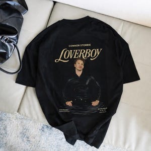 May include: Black t-shirt featuring a graphic of a man in water with the text "Loverboy" and "Connor Storrie". The shirt also includes the text "Ilya Rozanov" and "Heated Rivalry".