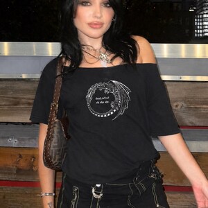 Op de afbeelding: Zwart off-the-shoulder T-shirt met een witte drakenafbeelding en de tekst "SenLinVu? oh she ate." Het shirt wordt gecombineerd met een zwarte rok met gele stiksels en een bruine handtas.