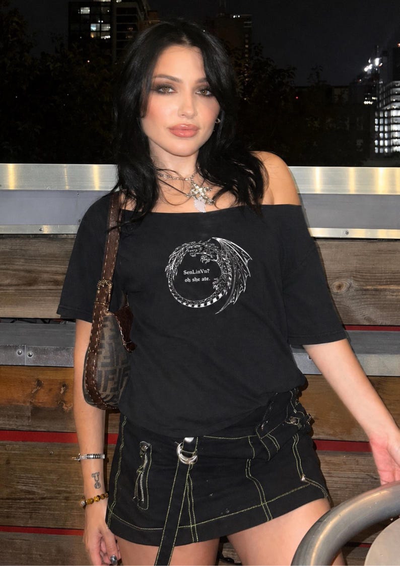 Op de afbeelding: Zwart off-the-shoulder T-shirt met een witte drakenafbeelding en de tekst "SenLinVu? oh she ate." Het shirt wordt gecombineerd met een zwarte rok met gele stiksels en een bruine handtas.
