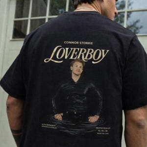 May include: Black t-shirt with the word "BLACK" in a black rectangle at the top. The shirt features the word "LOVERBOY" in a stylized font, the name "CONNOR STORRIE", and an image of a person in water. Text at the bottom reads "HEATED RIVALRY 10/01/26".