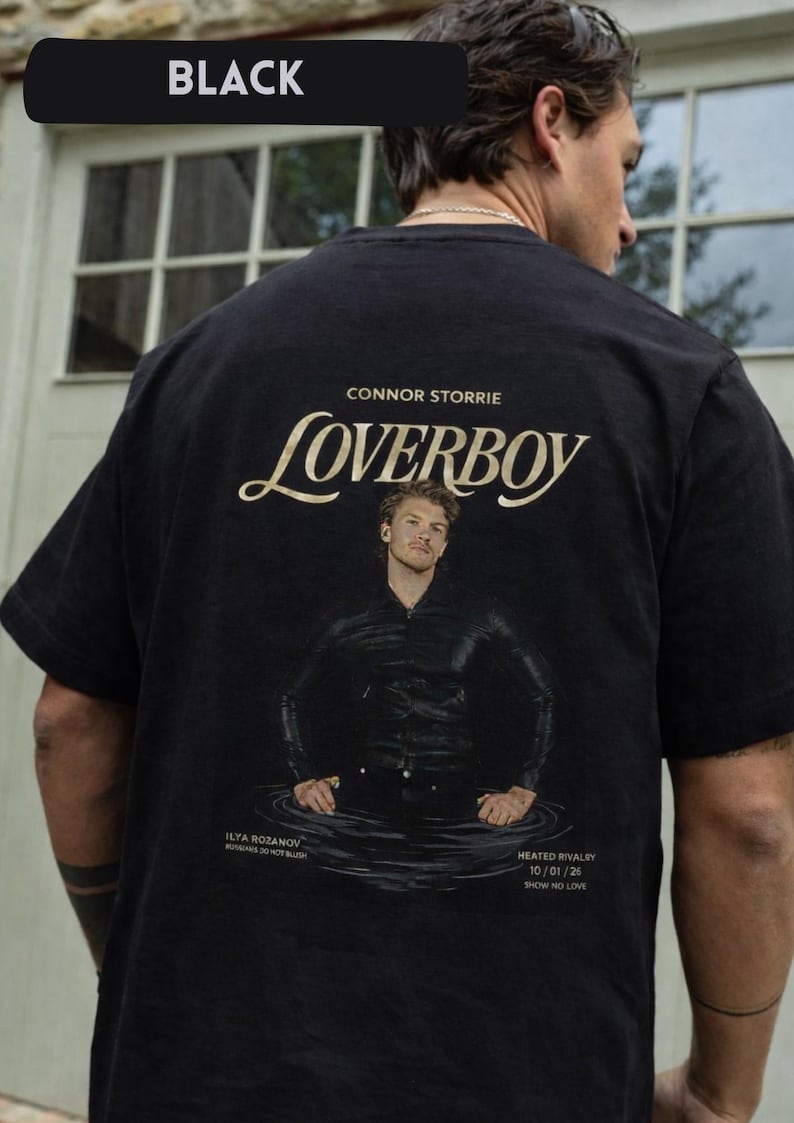 May include: Black t-shirt with the word "BLACK" in a black rectangle at the top. The shirt features the word "LOVERBOY" in a stylized font, the name "CONNOR STORRIE", and an image of a person in water. Text at the bottom reads "HEATED RIVALRY 10/01/26".