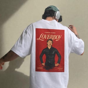 May include: White oversized t-shirt featuring a red graphic on the back with the text "CONNOR STORRIE Loverboy". The graphic shows a person in a black outfit standing in water. The shirt has a casual, relaxed fit.