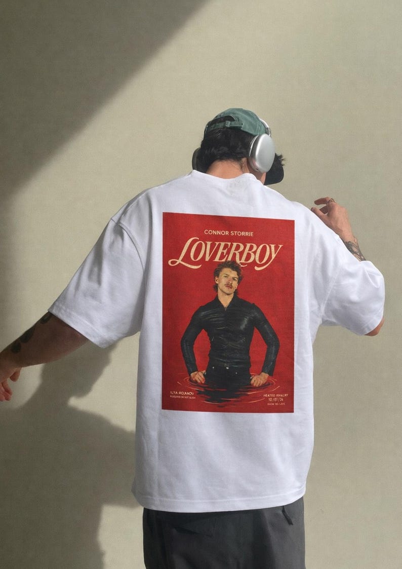 May include: White oversized t-shirt featuring a red graphic on the back with the text "CONNOR STORRIE Loverboy". The graphic shows a person in a black outfit standing in water. The shirt has a casual, relaxed fit.