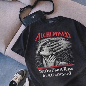 May include: Black graphic t-shirt with the word "ALCHEMISED" in red, featuring an illustration of a crying woman with hands covering her face. The text "You're Like A Rose In A Graveyard" is below the illustration. A black purse and sneakers are also in the image.