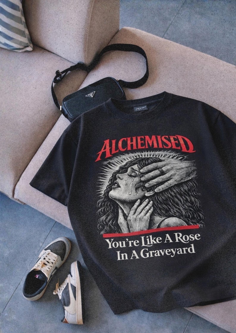 May include: Black graphic t-shirt with the word "ALCHEMISED" in red, featuring an illustration of a crying woman with hands covering her face. The text "You're Like A Rose In A Graveyard" is below the illustration. A black purse and sneakers are also in the image.