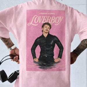 May include: A light pink t-shirt with a graphic on the back. The graphic is a pink magazine cover with the word "Loverboy" and a photo of a man in a black leather jacket.