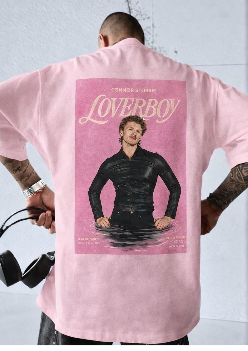 May include: A light pink t-shirt with a graphic on the back. The graphic is a pink magazine cover with the word "Loverboy" and a photo of a man in a black leather jacket.