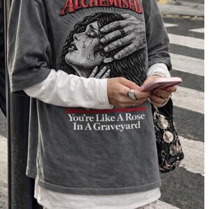 May include: Granite-colored graphic tee with the word "Alchemised" in red, featuring a distressed illustration of a crying woman. The text "You're Like A Rose In A Graveyard" is printed below the illustration.
