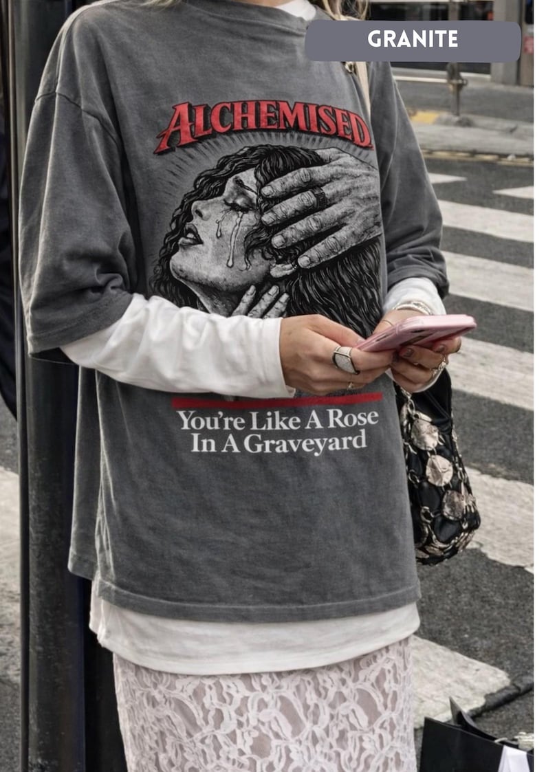 May include: Granite-colored graphic tee with the word "Alchemised" in red, featuring a distressed illustration of a crying woman. The text "You're Like A Rose In A Graveyard" is printed below the illustration.