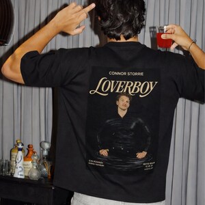 May include: Black t-shirt with a graphic featuring the word "Loverboy" in a stylized font, along with a portrait of a man. The shirt also includes the name "Connor Storrie" and other text. The person is wearing light-wash denim jeans.