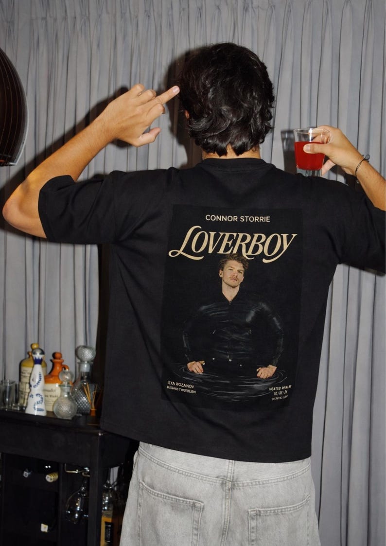 May include: Black t-shirt with a graphic featuring the word "Loverboy" in a stylized font, along with a portrait of a man. The shirt also includes the name "Connor Storrie" and other text. The person is wearing light-wash denim jeans.