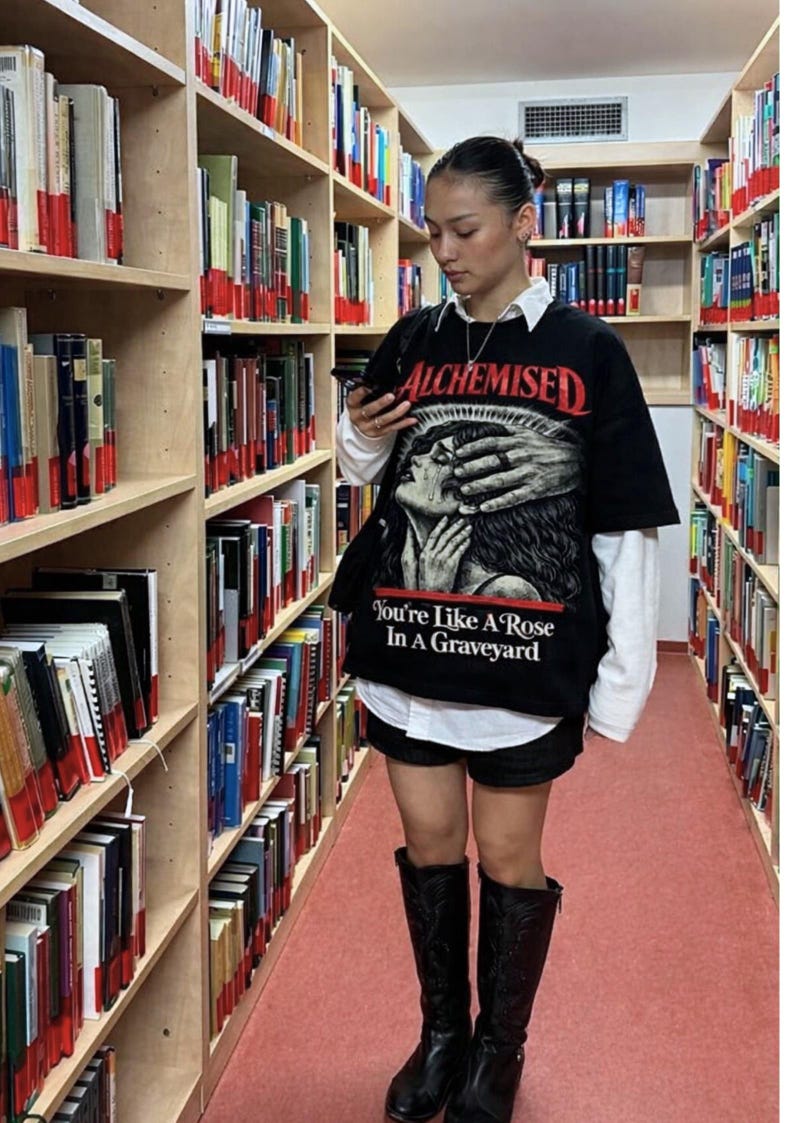 May include: Black graphic t-shirt with the word "ALCHEMISED" in red and a graphic of a person's face being covered by hands. The text "You're Like A Rose In A Graveyard" is below the graphic. The person is wearing black boots.
