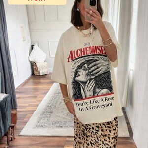 May include: Ivory-colored oversized t-shirt with a graphic print featuring the word "Alchemise" in red, and a distressed illustration of a crying woman. The text "You're Like A Rose In A Graveyard" is below the illustration. The model is wearing a leopard print skirt.
