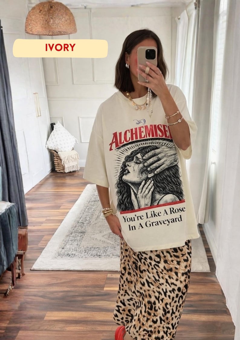 May include: Ivory-colored oversized t-shirt with a graphic print featuring the word "Alchemise" in red, and a distressed illustration of a crying woman. The text "You're Like A Rose In A Graveyard" is below the illustration. The model is wearing a leopard print skirt.
