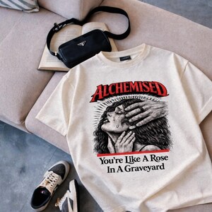 May include: Cream-colored t-shirt with the word "ALCHEMISED" in red, and the phrase "You're Like A Rose In A Graveyard". The graphic depicts a crying woman with hands over her face. A black purse, book, and sneakers are also visible.