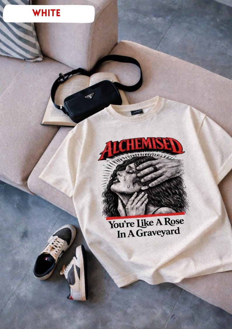 May include: Cream-colored t-shirt with the word "ALCHEMISED" in red, and the phrase "You're Like A Rose In A Graveyard". The graphic depicts a crying woman with hands over her face. A black purse, book, and sneakers are also visible.