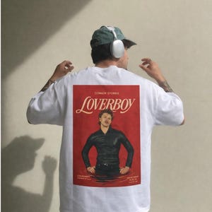 May include: White t-shirt with a red graphic featuring the word "LOVERBOY" and a man in a black outfit. The shirt also has the text "CONNOR STORRIE" and other text at the bottom. The model is wearing a green cap and headphones.