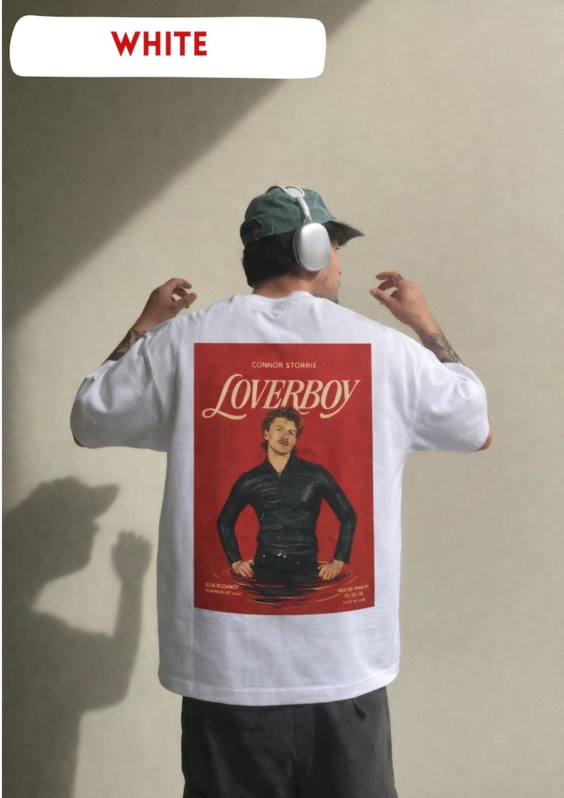 May include: White t-shirt with a red graphic featuring the word "LOVERBOY" and a man in a black outfit. The shirt also has the text "CONNOR STORRIE" and other text at the bottom. The model is wearing a green cap and headphones.