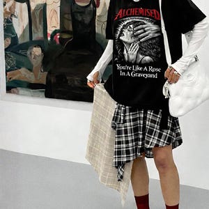 May include: Black graphic t-shirt with the text "ALCHEMISED You're Like A Rose In A Graveyard". The outfit includes a plaid skirt, white long-sleeve shirt, black sneakers, and a white shoulder bag. A beige scarf is held in the hand.