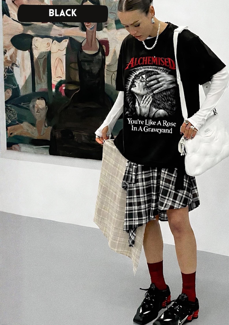 May include: Black graphic t-shirt with the text "ALCHEMISED You're Like A Rose In A Graveyard". The outfit includes a plaid skirt, white long-sleeve shirt, black sneakers, and a white shoulder bag. A beige scarf is held in the hand.