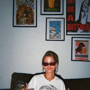Op de afbeelding: Een vrouw draagt een zonnebril en een wit t-shirt met een drakenafbeelding en de tekst "SenLinYu oh she ate." Verschillende ingelijste vintage posters hangen aan de muur achter haar.
