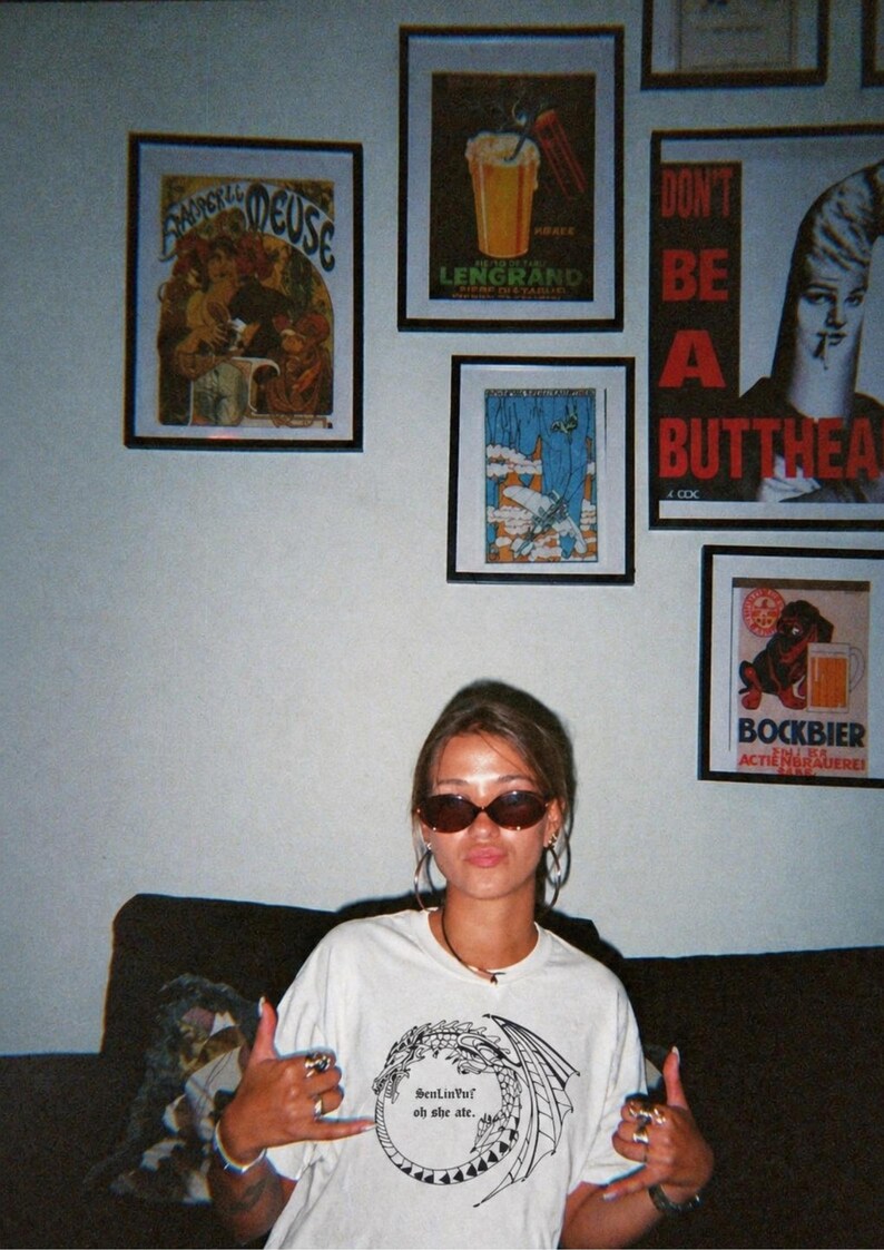 Op de afbeelding: Een vrouw draagt een zonnebril en een wit t-shirt met een drakenafbeelding en de tekst "SenLinYu oh she ate." Verschillende ingelijste vintage posters hangen aan de muur achter haar.
