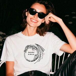 Op de afbeelding: Wit t-shirt met een zwart drakenontwerp en de tekst "SenLinYu? oh she ate." De persoon die het t-shirt draagt, draagt ook een zwarte zonnebril. Het woord "WHITE" staat in een witte rechthoek bovenaan.