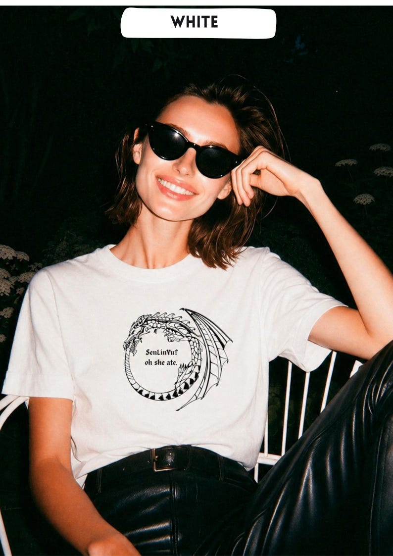Op de afbeelding: Wit t-shirt met een zwart drakenontwerp en de tekst "SenLinYu? oh she ate." De persoon die het t-shirt draagt, draagt ook een zwarte zonnebril. Het woord "WHITE" staat in een witte rechthoek bovenaan.