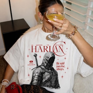May include: White t-shirt featuring a graphic of a hooded figure with a sword and the text "HARLAN". Additional text includes "The Destroyer, The Battle Ender, The Ancient Sword Wielder, He said you rise, always."