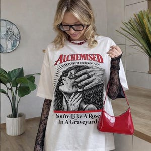 May include: White t-shirt with the word "ALCHEMISED" in red and a graphic of a crying woman with a hand over her face. The text "You're Like A Rose In A Graveyard" is below the graphic. A red patent leather handbag is also visible.