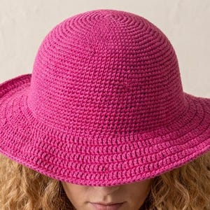 May include: A vibrant fuchsia pink crochet hat featuring a rounded crown and a wide, slightly downturned brim. The hat is constructed with a dense, uniform stitch pattern throughout, creating a textured surface that maintains its shape.
