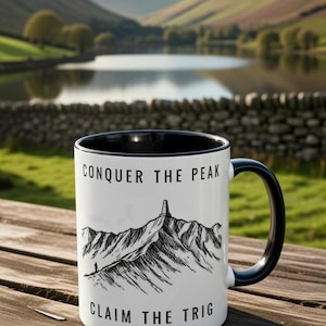 以下が含まれることがあります： 黒い内側とハンドルが付いた白いセラミックマグ。マグには、「CONQUER THE PEAK」と「CLAIM THE TRIG」というテキストが入った白黒の山のイラストが描かれています。マグは木製の表面に置かれています。