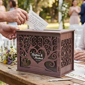 Op de afbeelding: Een houten trouwkaartendoos met een boomontwerp en de namen "Sophia & Mickael" en de datum "06/15/26" gegraveerd op de voorkant. Een persoon steekt een kaart met "Gefeliciteerd!" in de gleuf.