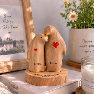 Può includere: Due statuette di pinguini in legno marrone chiaro poggiano su una base circolare in legno. Il pinguino più piccolo ha un cuore rosso e il nome Emma, mentre quello più grande ha un cuore rosso e il nome Liam. La base riporta Happy Arniversary 4/21/2021.