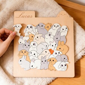Könnte beinhalten: Holzpuzzle mit Cartoon-Hasen in Weiß, Grau und Beige. Das Puzzle hat einen Lasche mit dem eingravierten Namen "Lucas". Eine Hand hält ein Hasenstück. Das Puzzle ist aus Holz.