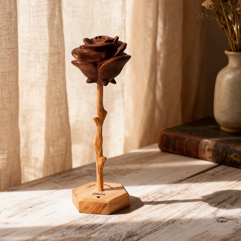 K&ouml;nnte beinhalten: Eine geschnitzte Holzrosenskulptur mit einem dunkelbraunen Bl&uuml;tenkopf und einem hellbraunen Stiel. Die Rose ist auf einem sechseckigen Holzsockel montiert, der den eingravierten Text "Love forever" und den Namen "Olivia" mit einem kleinen Herzsymbol zeigt.