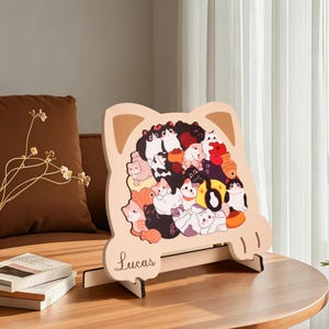 Könnte beinhalten: Dekoratives Holzstück in Katzenform mit einer Sammlung von Cartoon-Katzen in verschiedenen Farben. Das Stück hat einen hellbraunen Hintergrund mit dem Namen "Lucas" in Schreibschrift. Es steht auf einer Holzstaffelei.