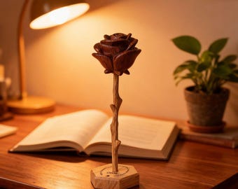 Peut inclure: Une sculpture de rose en bois faite à la main, avec une tête de fleur marron foncé et une tige marron clair. La rose est posée sur une base hexagonale en bois. La sculpture est exposée sur un bureau en bois avec un livre ouvert et une lampe de bureau.