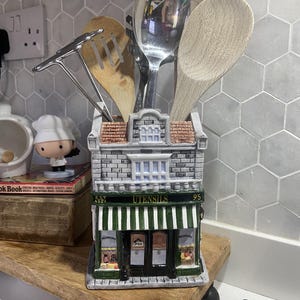 Może przedstawiać: Ceramiczny pojemnik na przybory kuchenne w kształcie budynku sklepu z zielono-białą markizą, napisem UTENSILS i ceglaną fasadą. W pojemniku znajduje się metalowa łyżka, metalowe narzędzie oraz dwie drewniane łyżki.