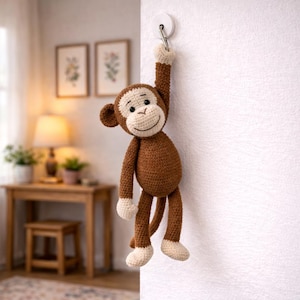 以下が含まれることがあります： 茶色とクリーム色の色合いの、かぎ針編みで作られた猿のおもちゃ。白いフックに吊り下げられています。猿は笑顔で、長い手足があり、吊り下げられるように設計されています。手作りの玩具です。