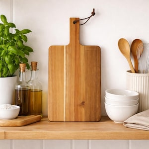Puede incluir: Tabla de cortar rectangular de madera clara, con asa y lazo de cuero. Se muestra en una estantería de madera junto con otros artículos de cocina, como cuencos, utensilios y una planta de albahaca.