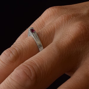 Może przedstawiać: Srebrny pierścionek z małym, okrągłym, czerwonym kamieniem szlachetnym w spiczastym wzorze. Pierścionek ma teksturowaną powierzchnię z małymi, równomiernie rozmieszczonymi kropkami. Pierścionek jest na palcu na ciemnym tle.