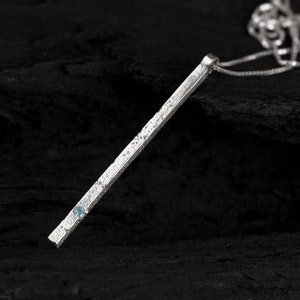 以下が含まれることがあります： 小さな青い宝石が付いた、長く、質感のあるバーペンダントが付いたシルバーネックレス。ネックレスは黒い背景の上にあります。