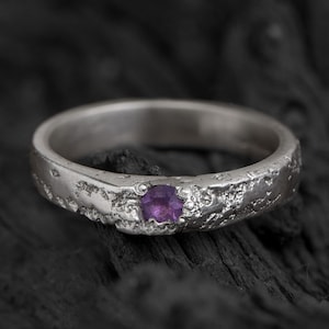Könnte beinhalten: Ein silberner Ring mit einer strukturierten Band und einem kleinen violetten Edelstein in der Mitte.