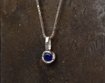 Op de afbeelding: Een zilveren ketting met een klein hanger met een donkerblauwe edelsteen.
