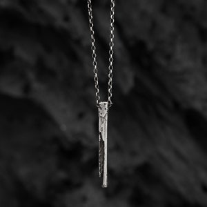 Puede incluir: Un collar de plata con un colgante largo, delgado y texturizado. El colgante es un dise&ntilde;o simple y minimalista.
