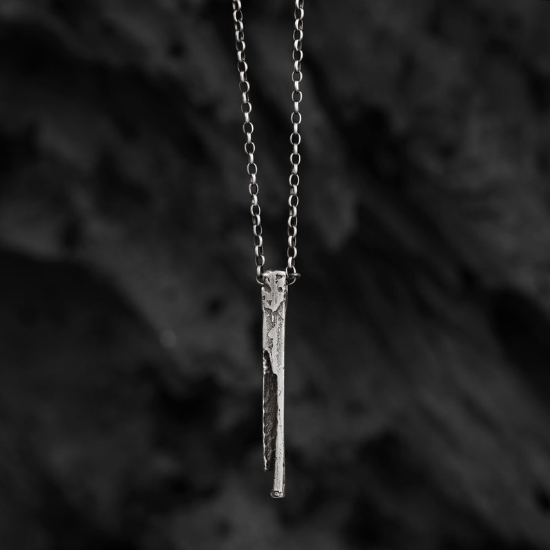 Puede incluir: Un collar de plata con un colgante largo, delgado y texturizado. El colgante es un dise&ntilde;o simple y minimalista.