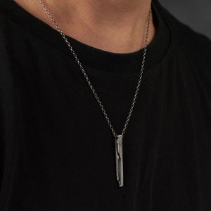 Puede incluir: Un collar de cadena de plata con un colgante largo, delgado y texturizado. El colgante est&aacute; hecho de metal y tiene un aspecto &aacute;spero y org&aacute;nico.
